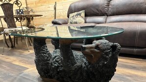 Un tavolino da caffè con piano in vetro e base a forma di orso, un divano in pelle e un cuscino decorativo in un soggiorno.
