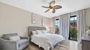 Une chambre à coucher avec un lit, un fauteuil gris, un ventilateur de plafond et une fenêtre avec des rideaux.