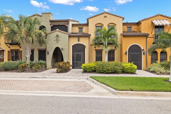 A row of Mediterranean-style buildings with arched doorways, palm trees, and a paved driveway. - Beautiful Townhouse in Paseo (Fort Myers)