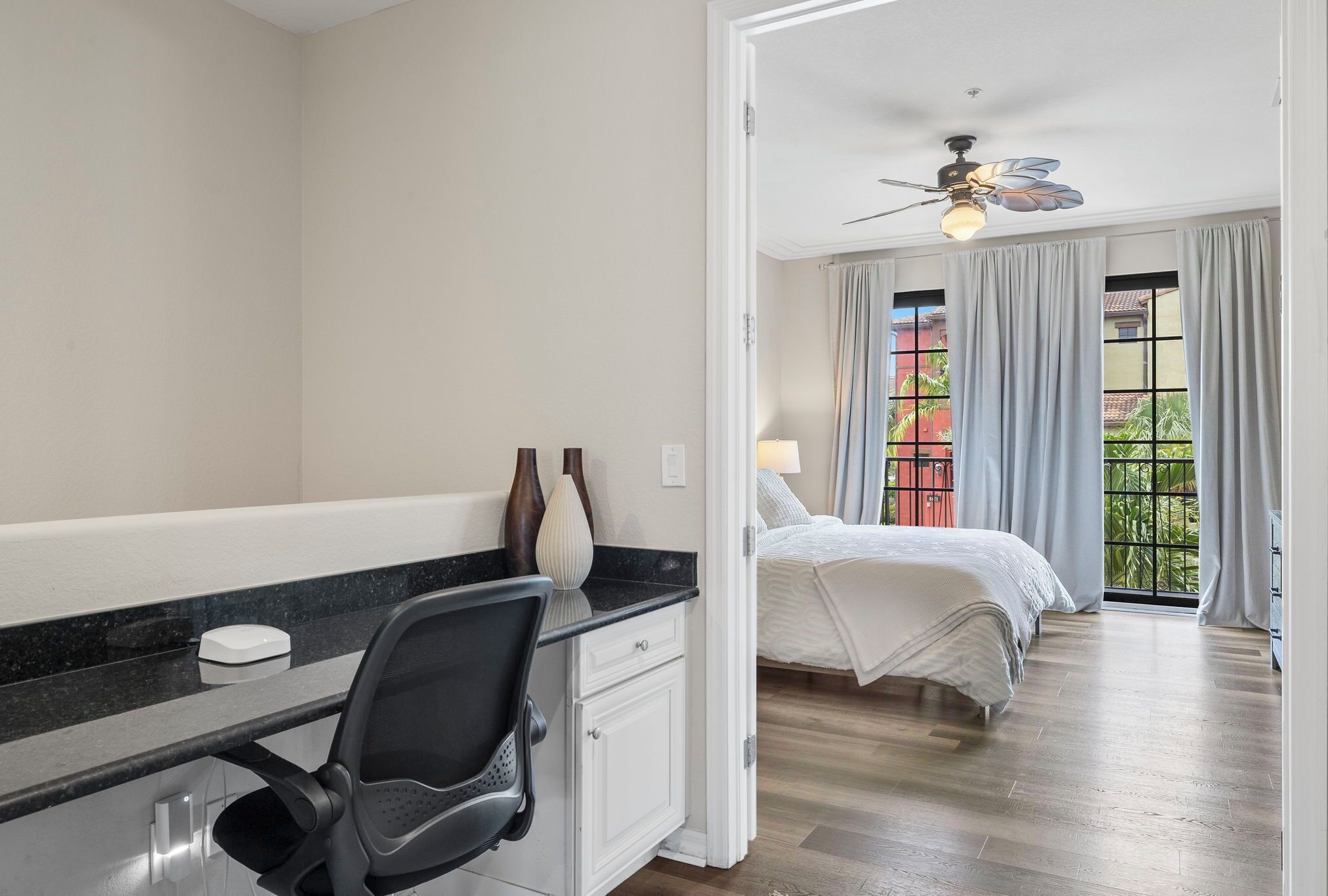 A bedroom with a bed, a desk with a chair, a ceiling fan, and a view of the outside through a window.