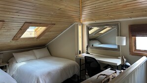 A bedroom with a sloped ceiling, a bed, a desk, a chair, a lamp, and a mirror.