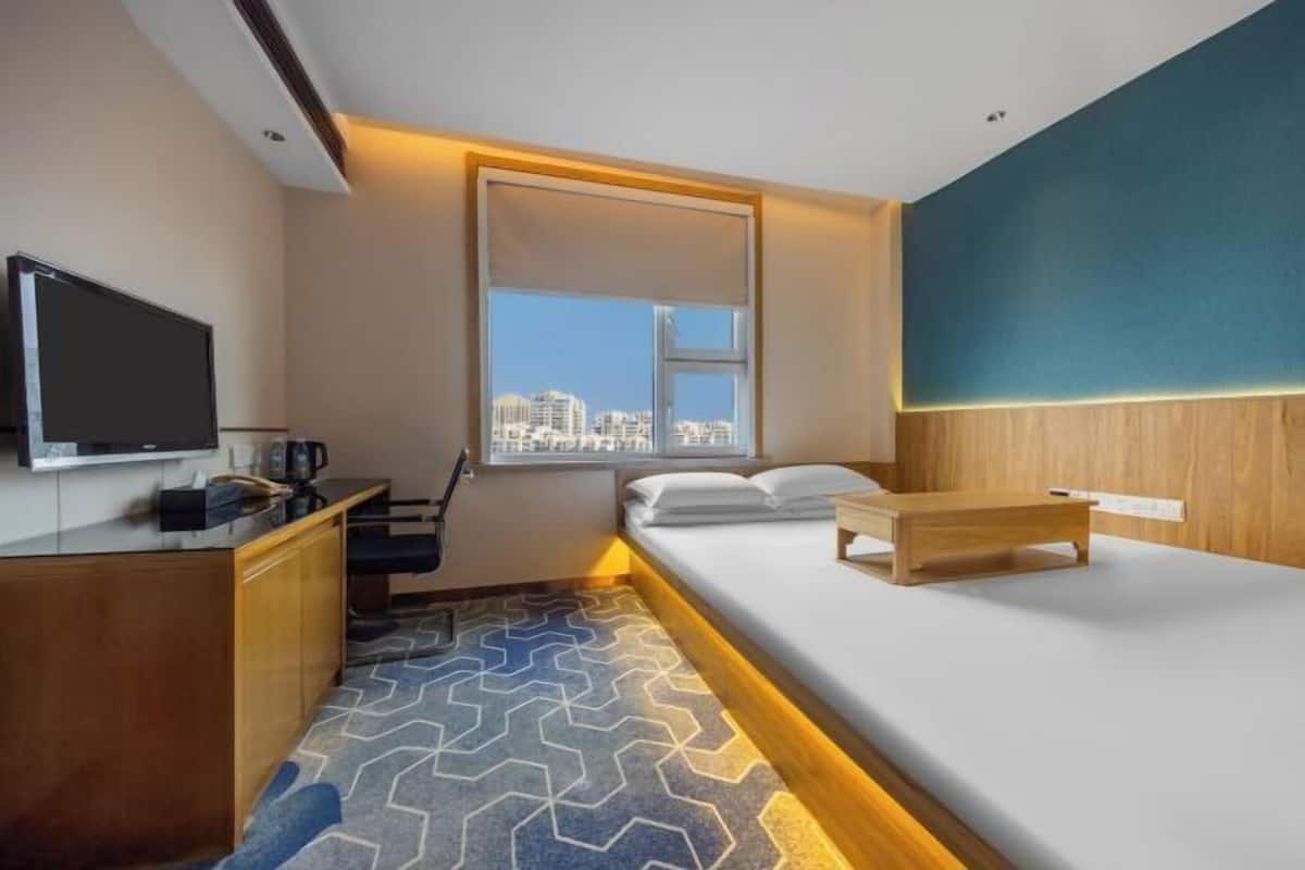 A hotel room with a bed, a desk with a flat-screen TV, a coffee maker, and a window with a city view.
