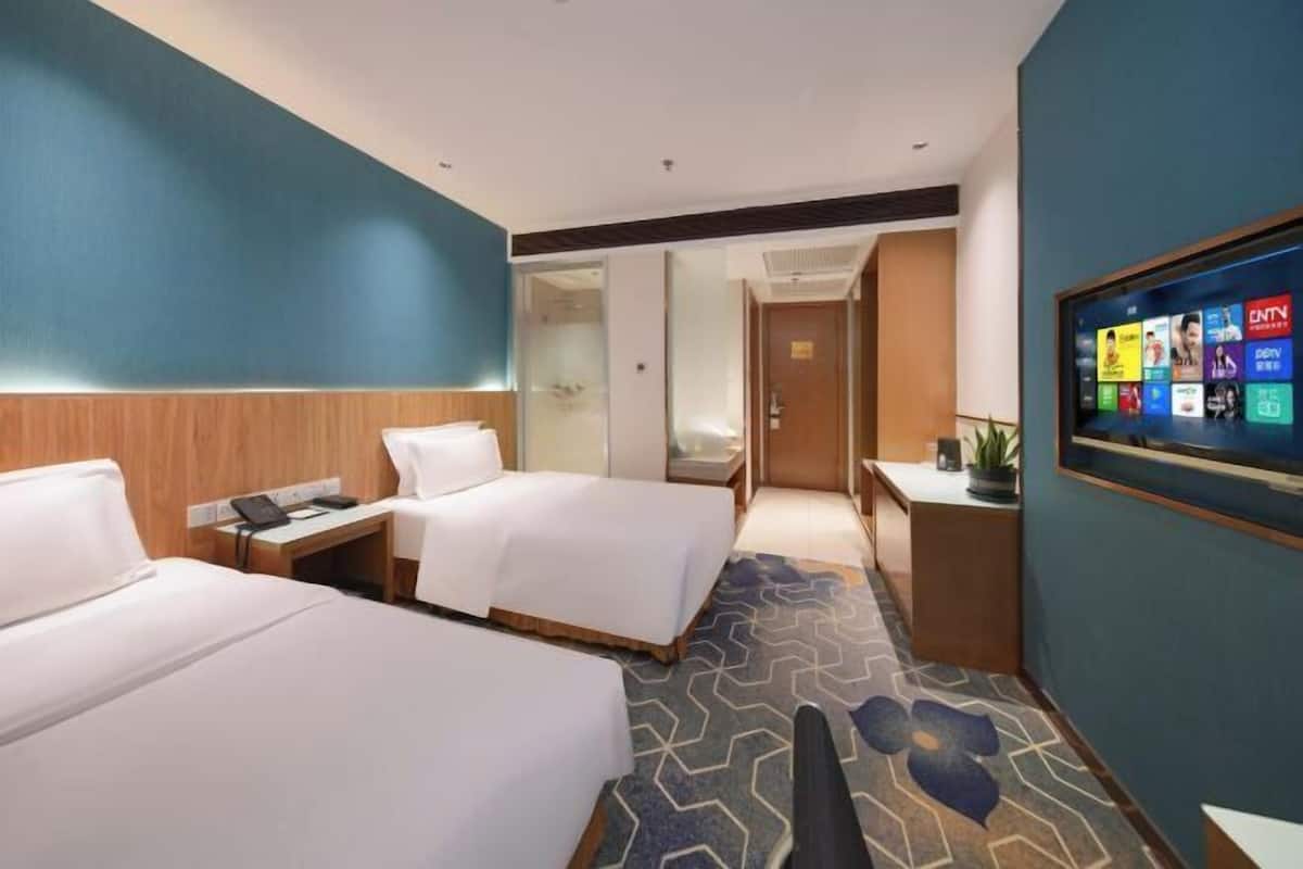 A hotel room with two beds, a desk with a computer, a TV displaying channels, and a patterned carpet.