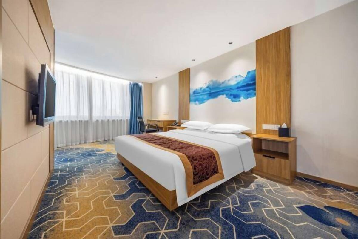 A hotel room with a bed, a flat-screen TV, a desk, a chair, a nightstand, a lamp, and a painting on the wall.