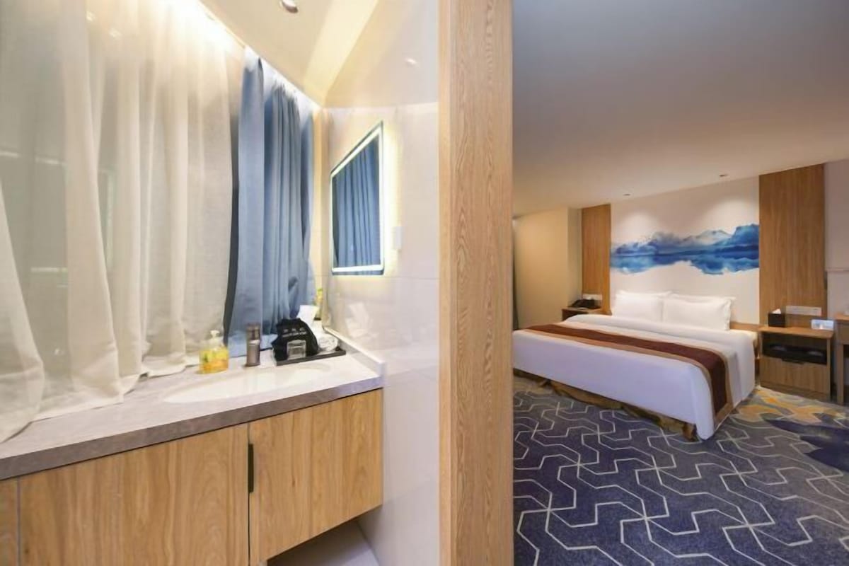 A hotel room with a bed, a bedside table, a nightstand, a mirror, a window with curtains, and a wall with a blue abstract painting.