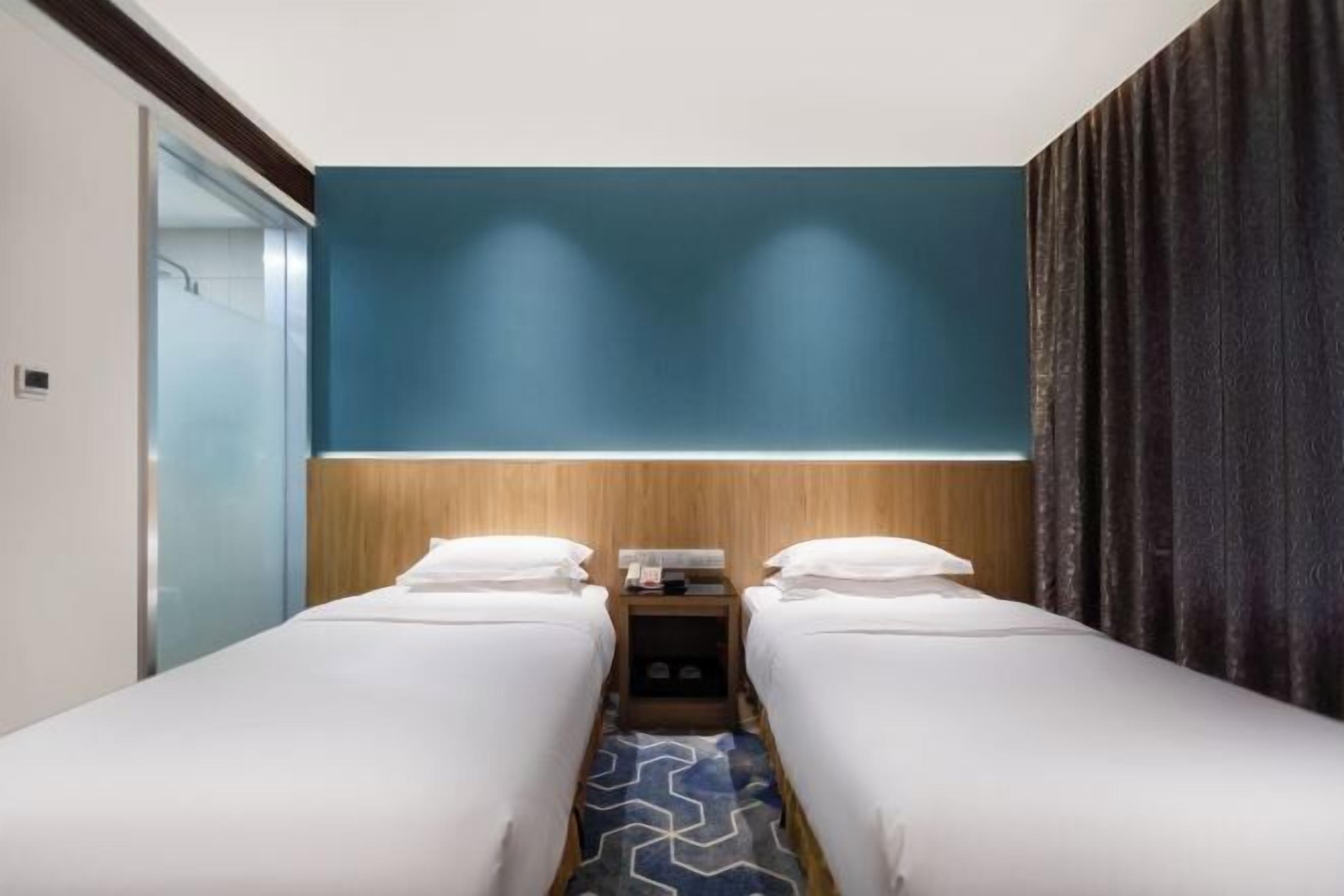 Habitación de hotel con dos camas individuales, cabecera de madera, mesita de noche pequeña con lámpara y una pared azul decorativa.