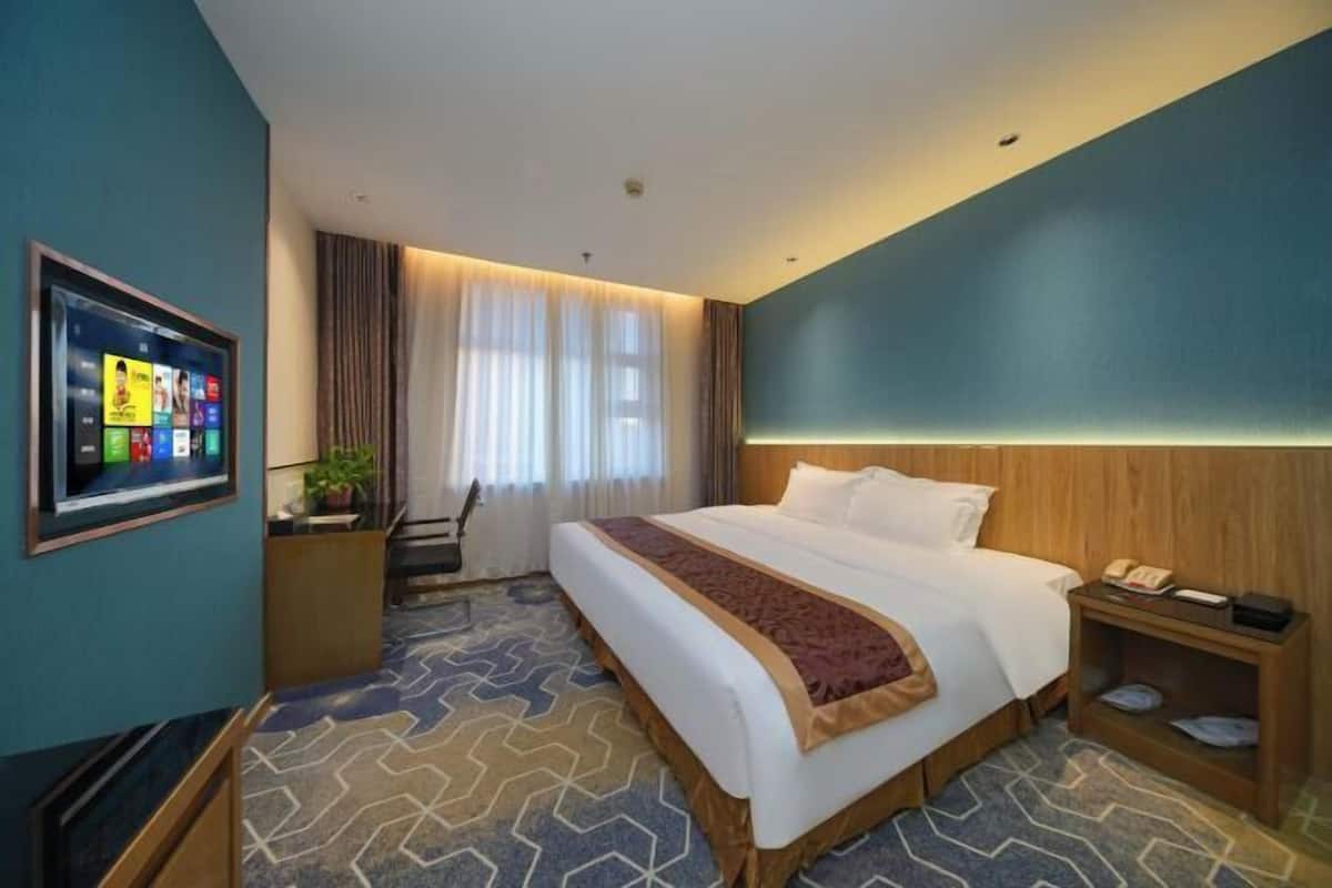 A hotel room with a large bed, a desk with a computer, a TV mounted on the wall, and a window with curtains.