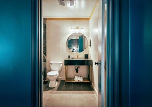 A bathroom with a white toilet, a black sink with a gold faucet, and a mirror with lights above it. - The Fainting Couch Boutique Hotel (Cobb)