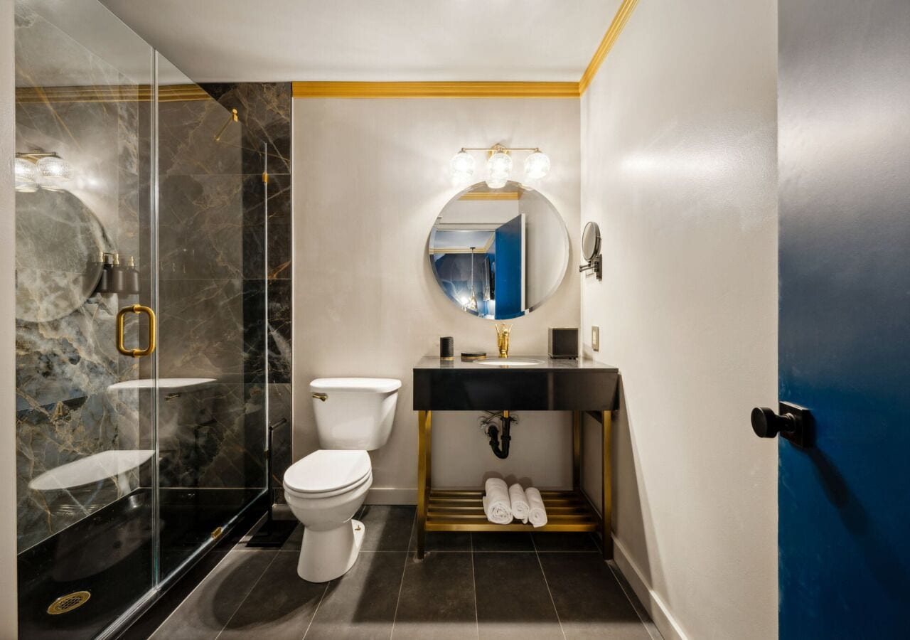A modern bathroom with a glass shower enclosure, a toilet, a sink with a gold base, and a mirror with lights.