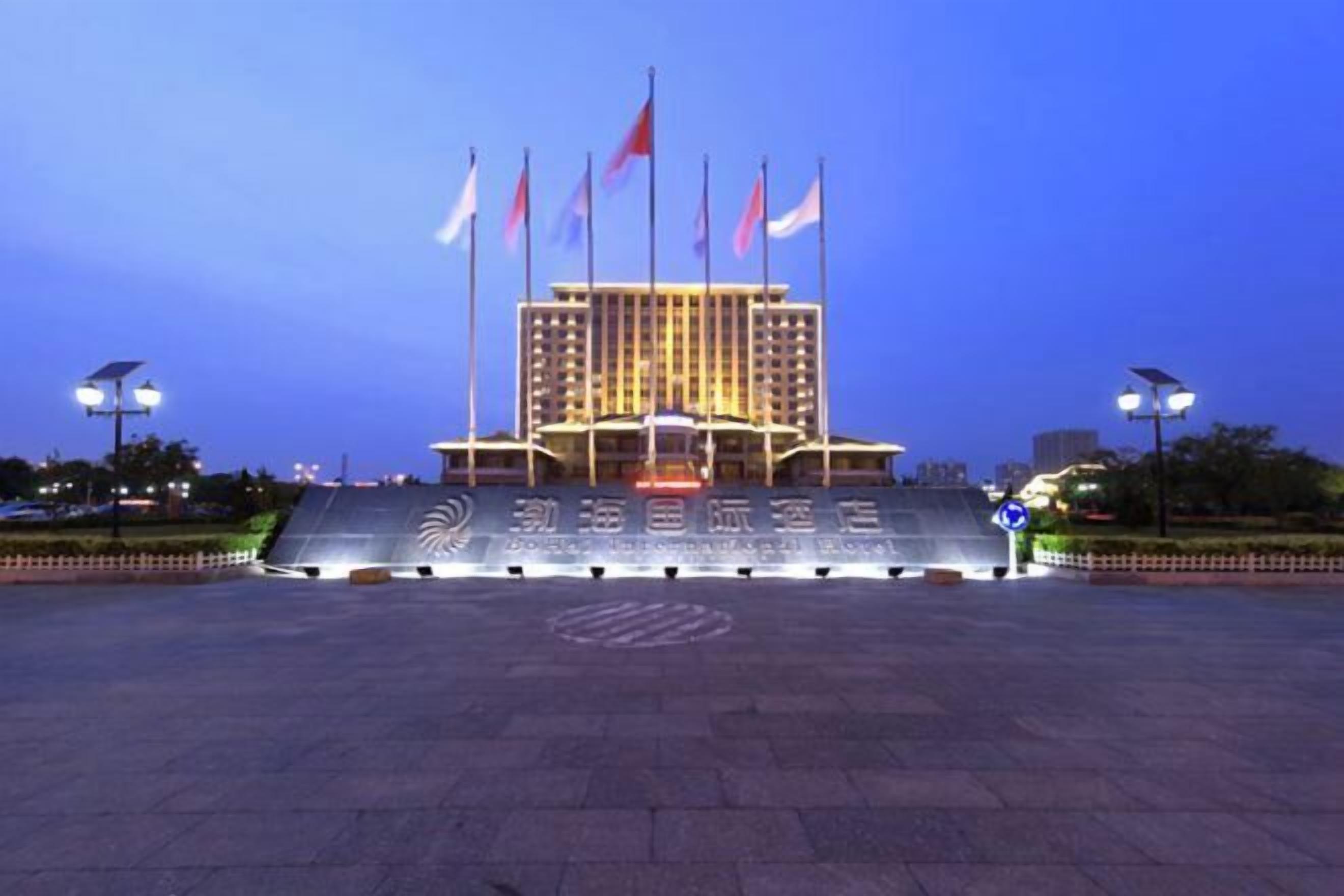 A large building with multiple flags, a paved entrance area, and a well-lit facade.