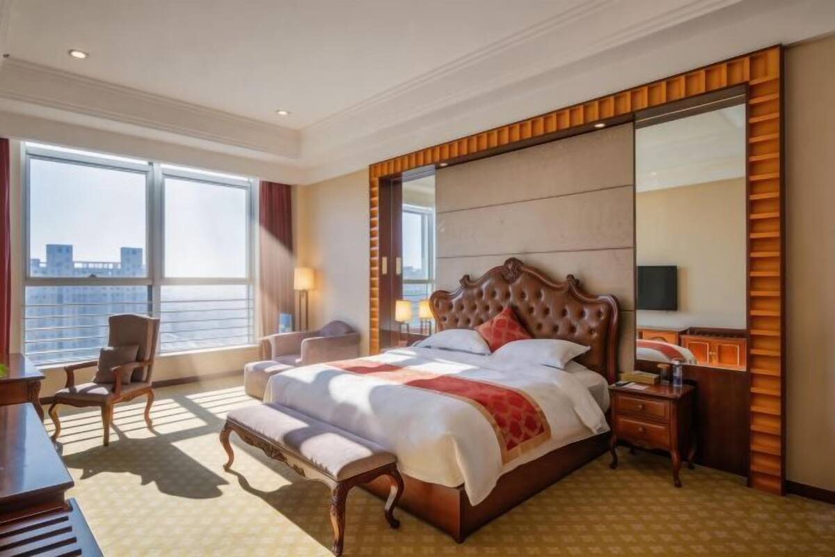 Ein Hotelzimmer mit einem großen Bett, einem Schreibtisch, einem Sessel und einem großen Spiegel, mit Blick auf die Stadt.