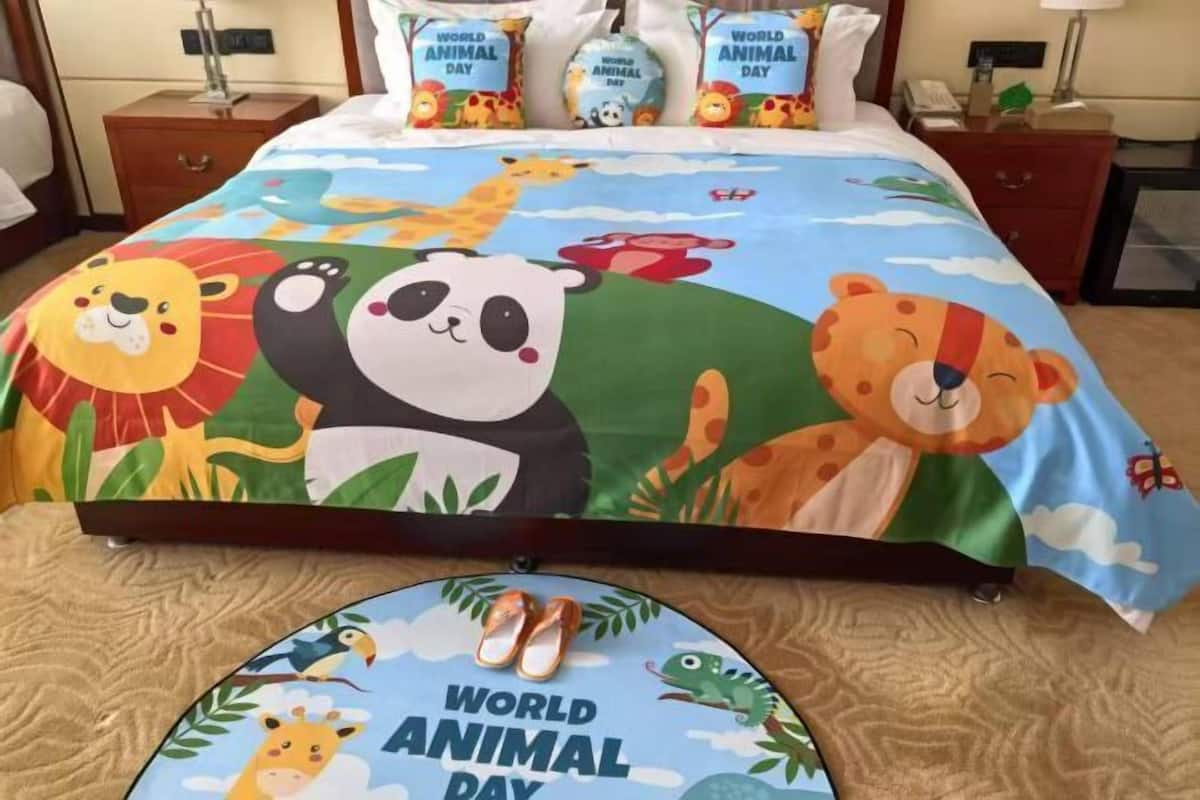 Ein Hotelzimmer mit einem Bett, ausgestattet mit einem farbenfrohen Bettbezug und Kissen mit Tiermotiven, einem runden Teppich mit dem Aufdruck 'World Animal Day' und einem Nachttisch mit Lampe und Telefon.