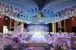 Un salón de banquetes con escenario, sillas blancas y decoración floral.