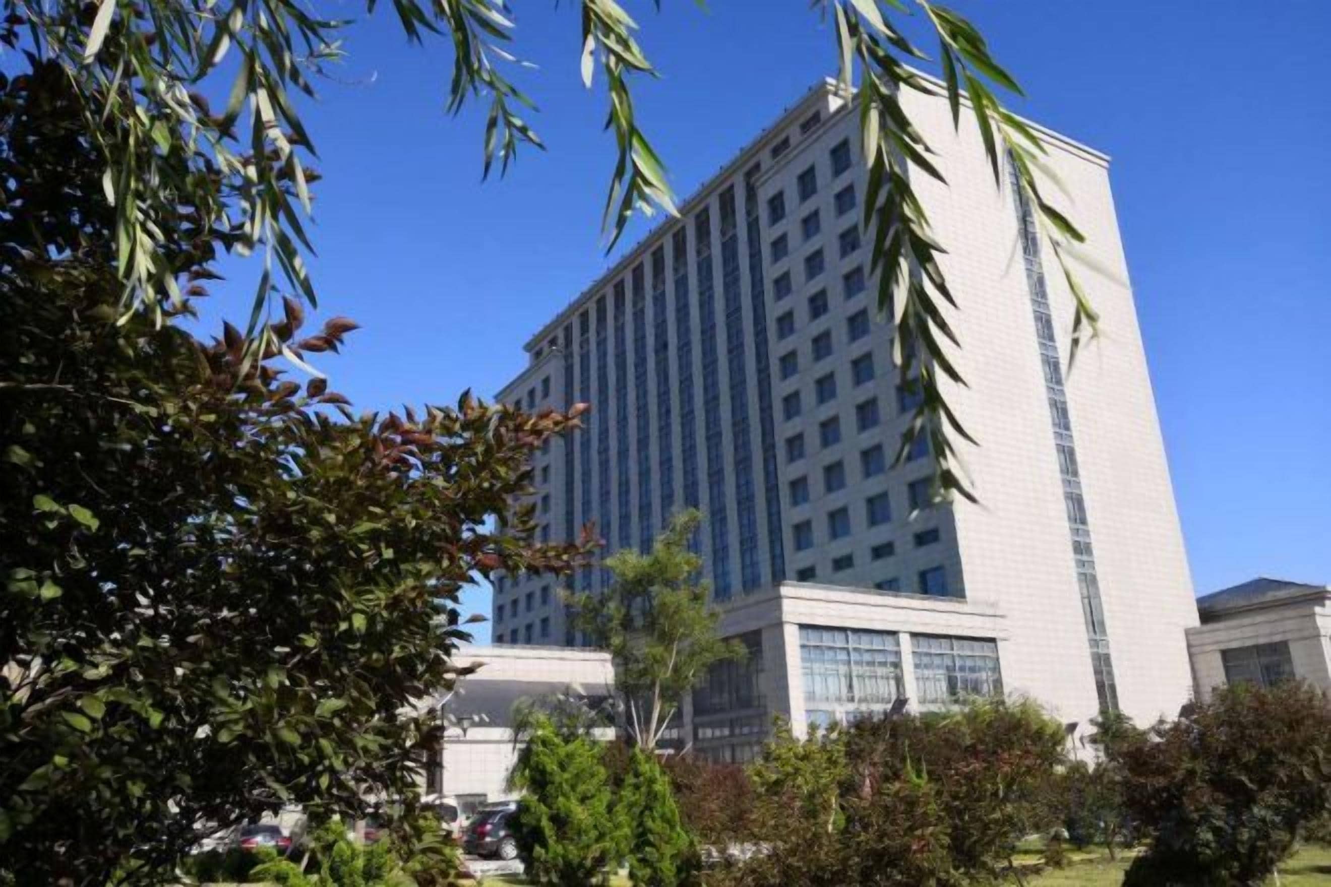 A tall, modern building with a glass facade, surrounded by greenery and a clear blue sky.