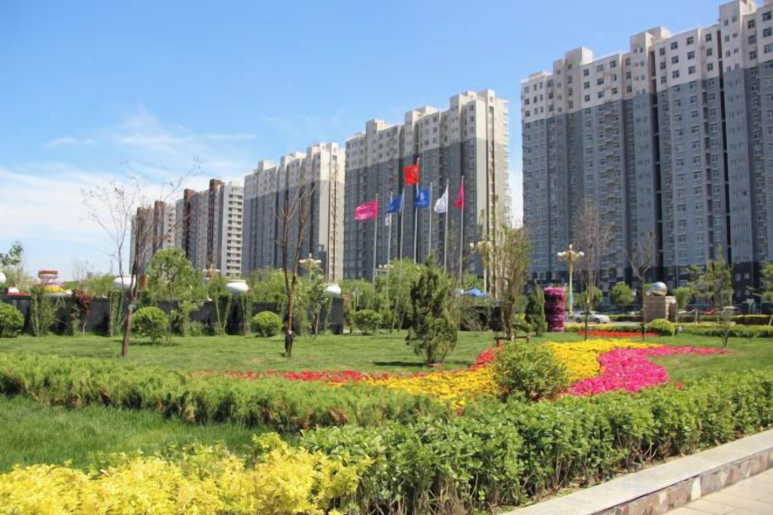Zona residencial con edificios altos, un jardín bien cuidado con flores coloridas y un cielo despejado.