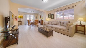 Un salón amplio con sofá, reposapiés y una mesa de centro de vidrio. Se observa un comedor con sillas y una mesa, y una cocina al fondo.