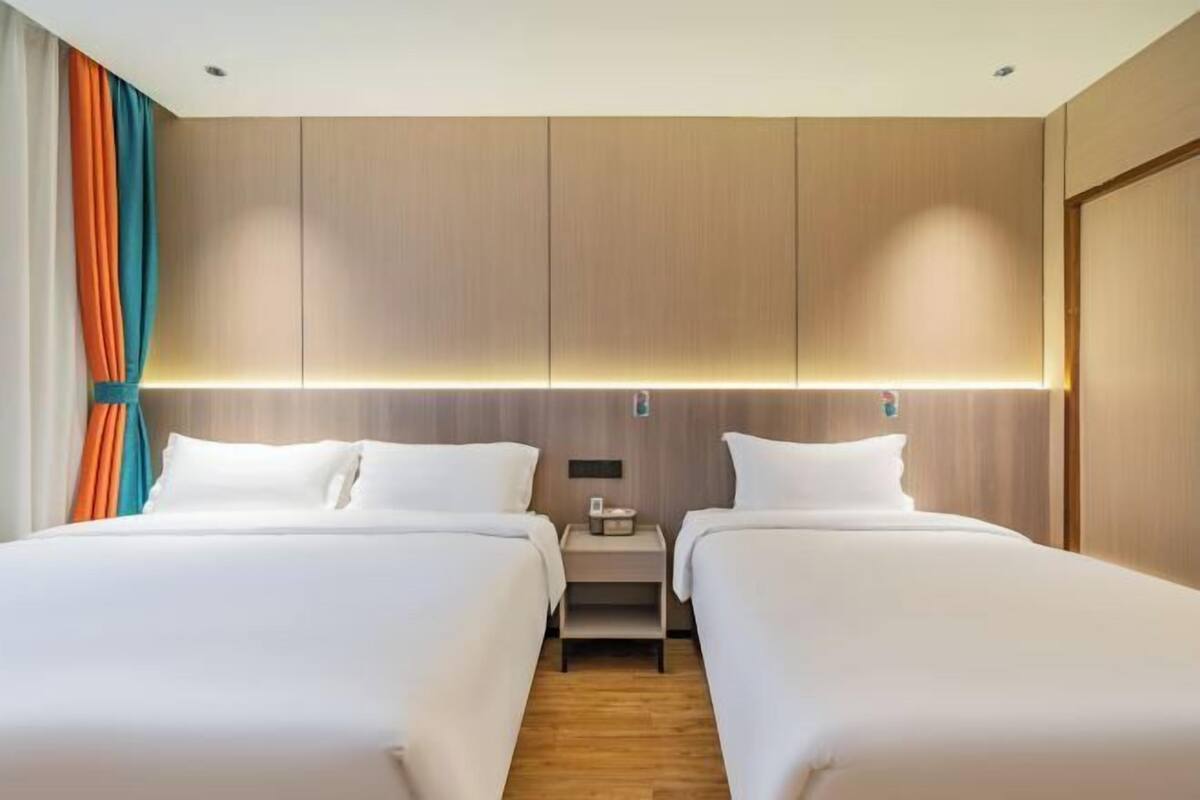 Habitación de hotel con dos camas, cabecero de madera, mesita de noche con lámpara y luz empotrada en la pared.