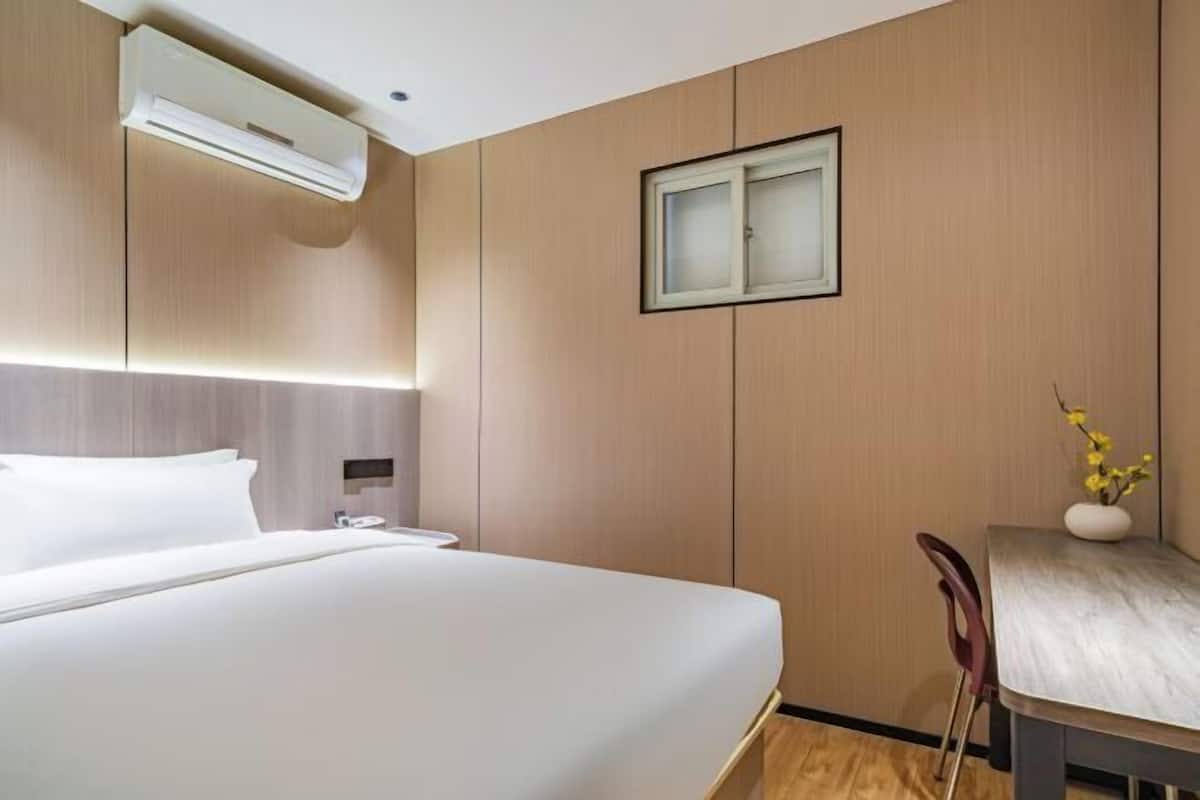 Habitación de hotel con cama, mesita de noche, lámpara y ventana pequeña.