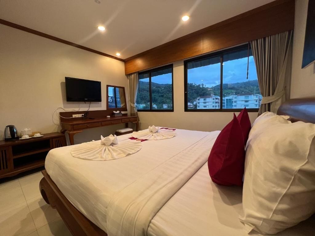Habitación de hotel con cama, televisión, escritorio con silla y ventanas amplias con vista a edificios y colinas.