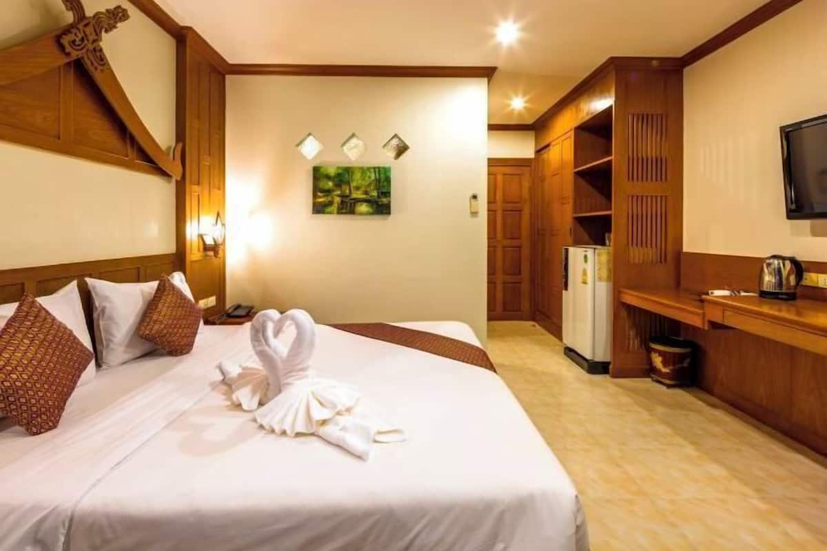 A hotel room with a neatly made bed, a wooden headboard, a wall-mounted TV, and a small kitchenette area.