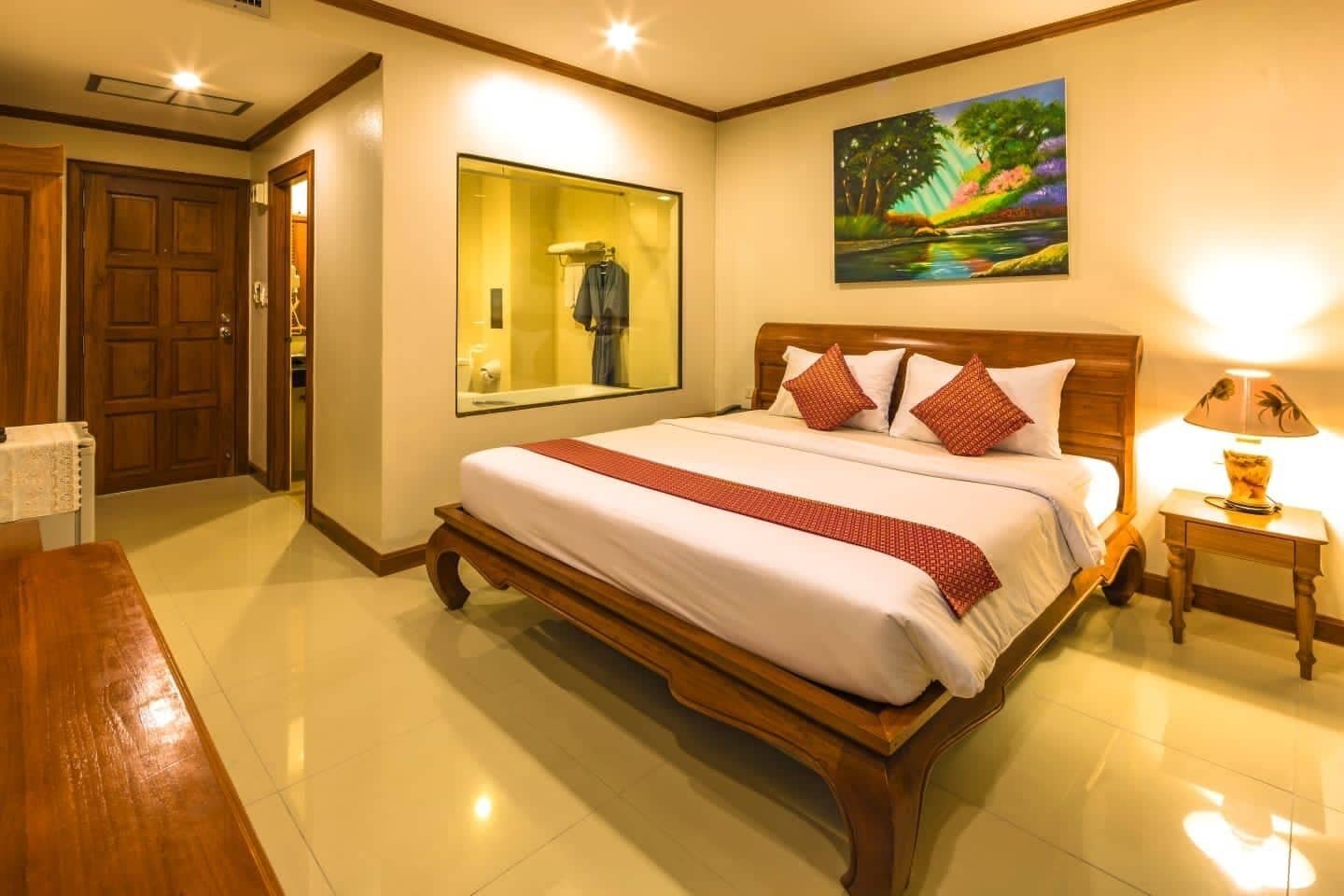 A hotel room with a large bed, a wooden headboard, a painting on the wall, a mirror, and a bedside table with a lamp.