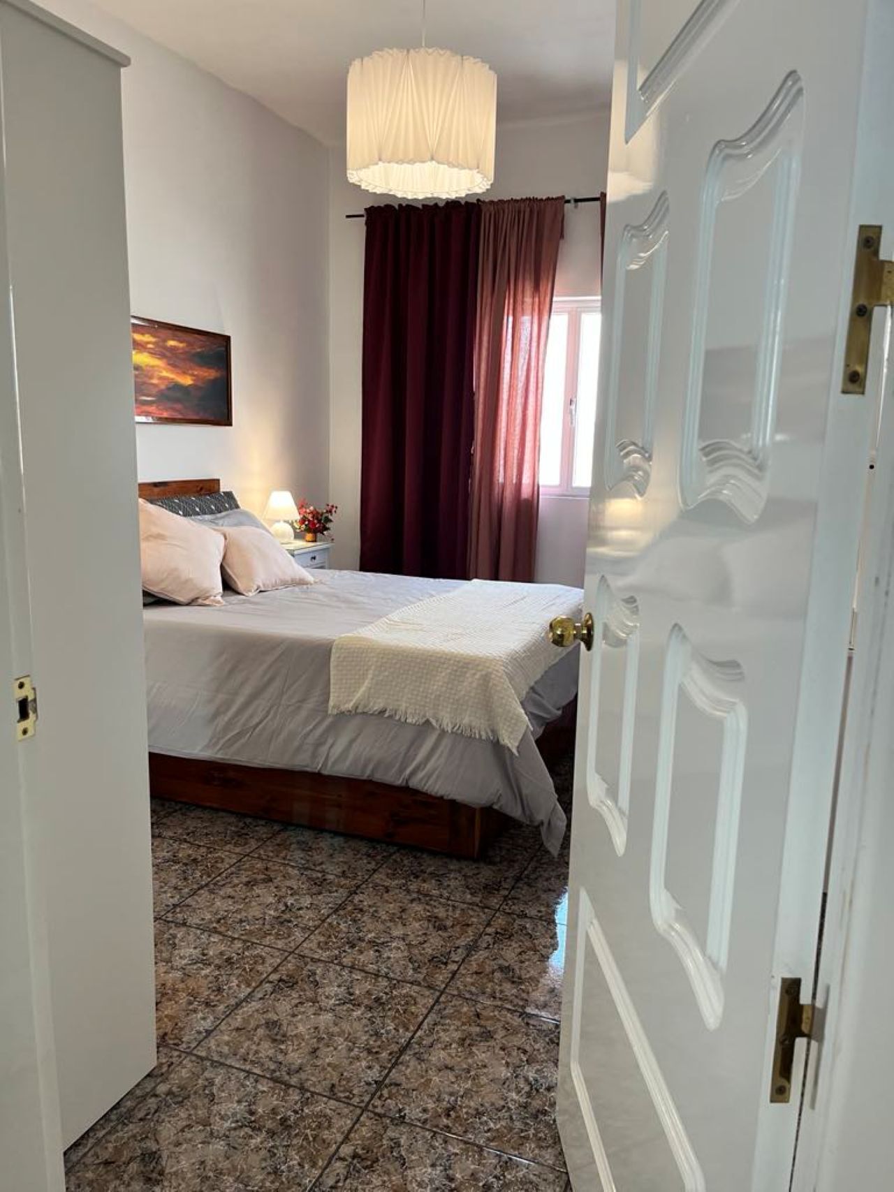 A bedroom with a bed, a nightstand, a lamp, a painting on the wall, and a window with curtains.