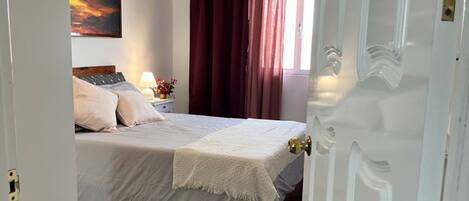 A bedroom with a bed, a nightstand, a lamp, a painting on the wall, and a window with curtains.