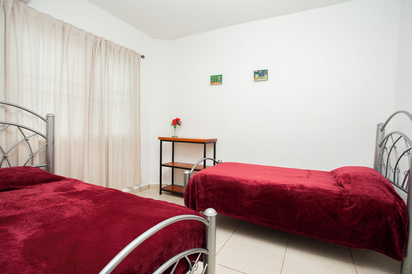 A room with two metal beds, a small wooden table with a vase of flowers, and two small wall decorations.