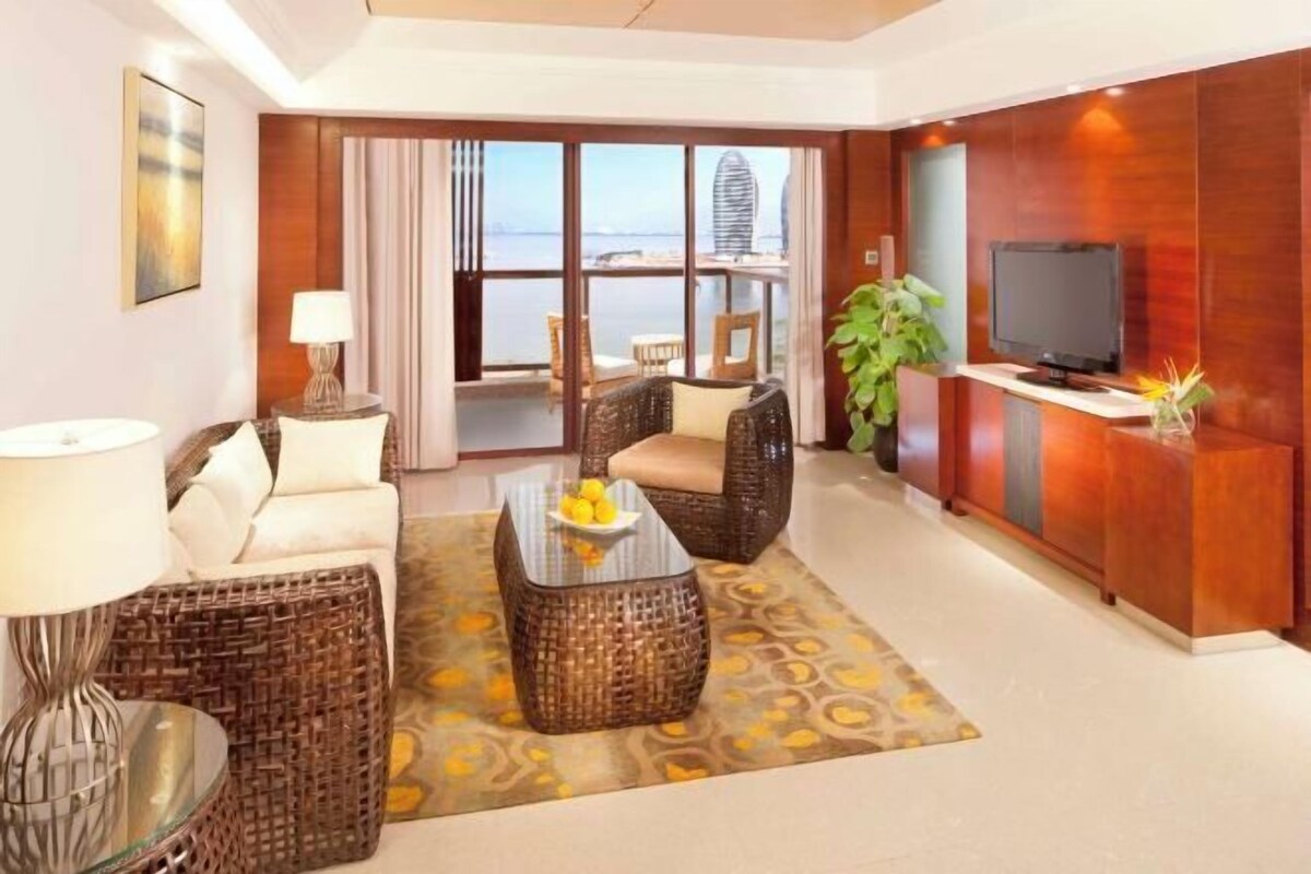 A hotel room with a sofa, a coffee table with fruit, a TV, and a balcony with a view of the sea.