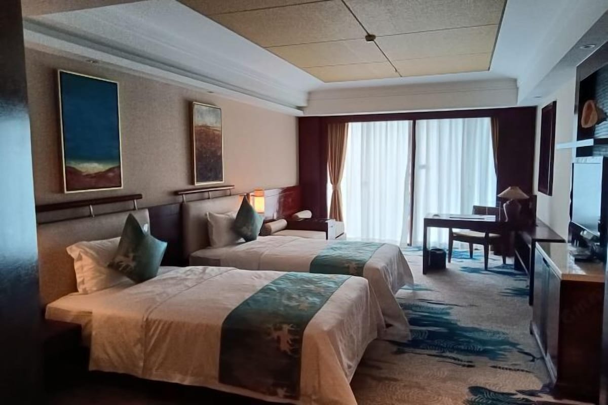 A hotel room with two beds, a desk, and a chair. There are paintings on the walls and a large window with curtains.