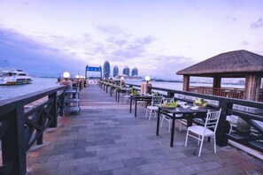 A waterfront restaurant with outdoor seating, a thatched-roof structure, and a view of the city skyline.