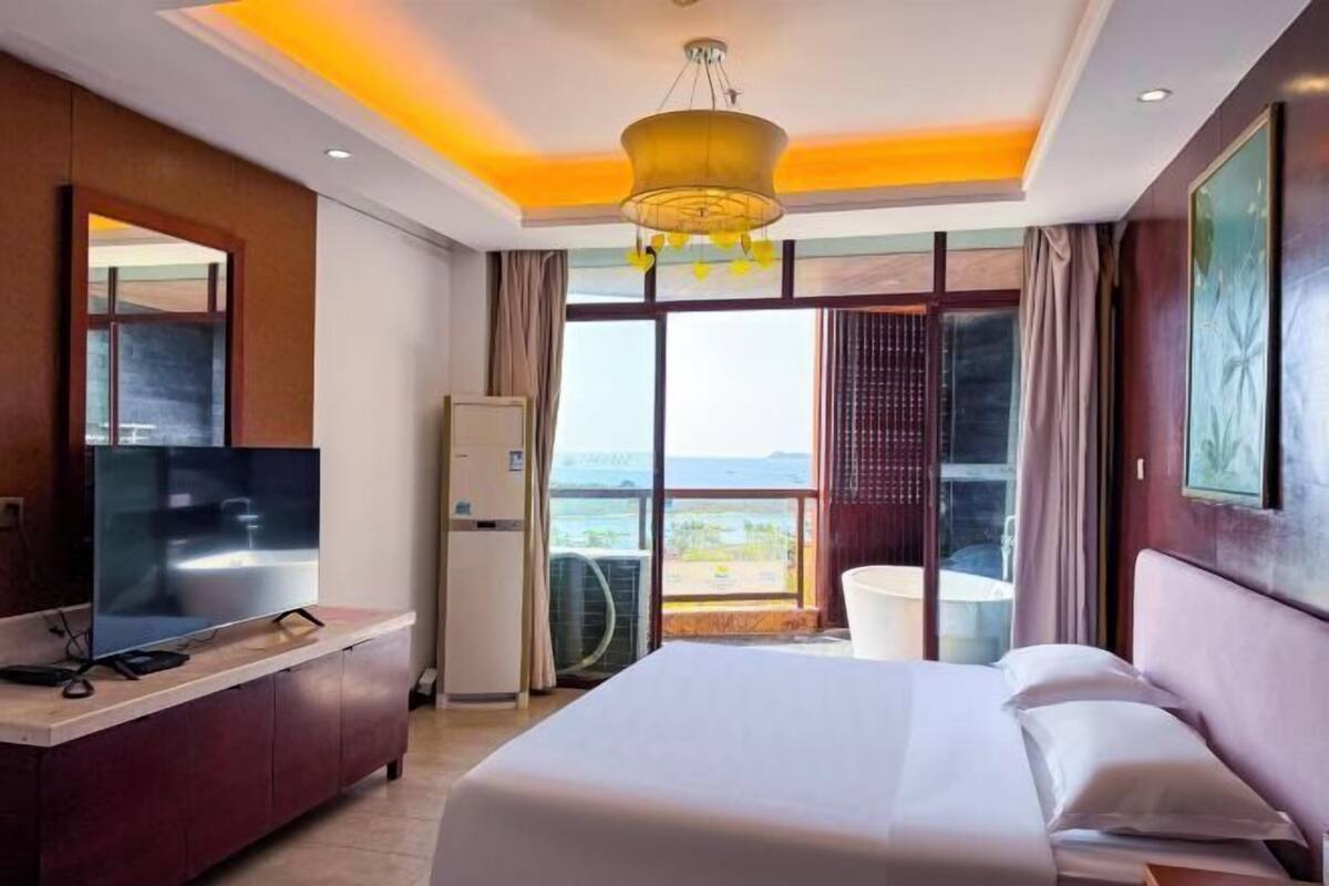 A hotel room with a bed, a TV, a minibar, a window with a view, and a ceiling light.