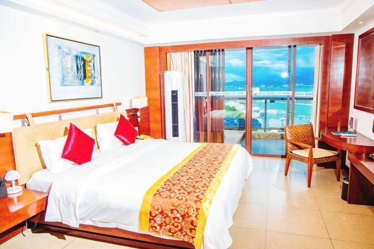 A hotel room with a bed, a desk, a chair, and a large window with a view of the sea.