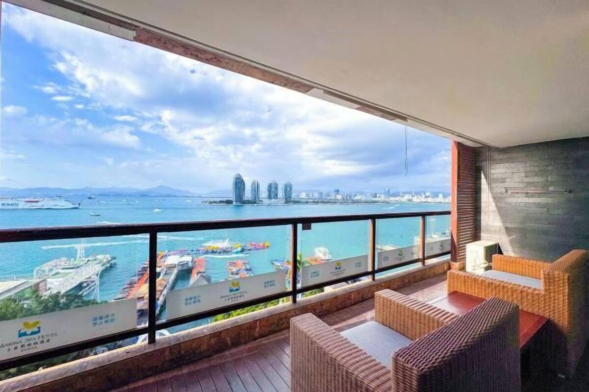 A balcony with wicker furniture, a view of a harbor with ships, and a city skyline in the distance.
