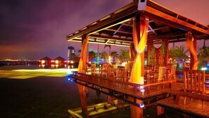 Un espace extérieur aménagé pour les repas, avec des tables et des chaises en bois, un coin salon abrité et une piscine qui reflète les lumières.