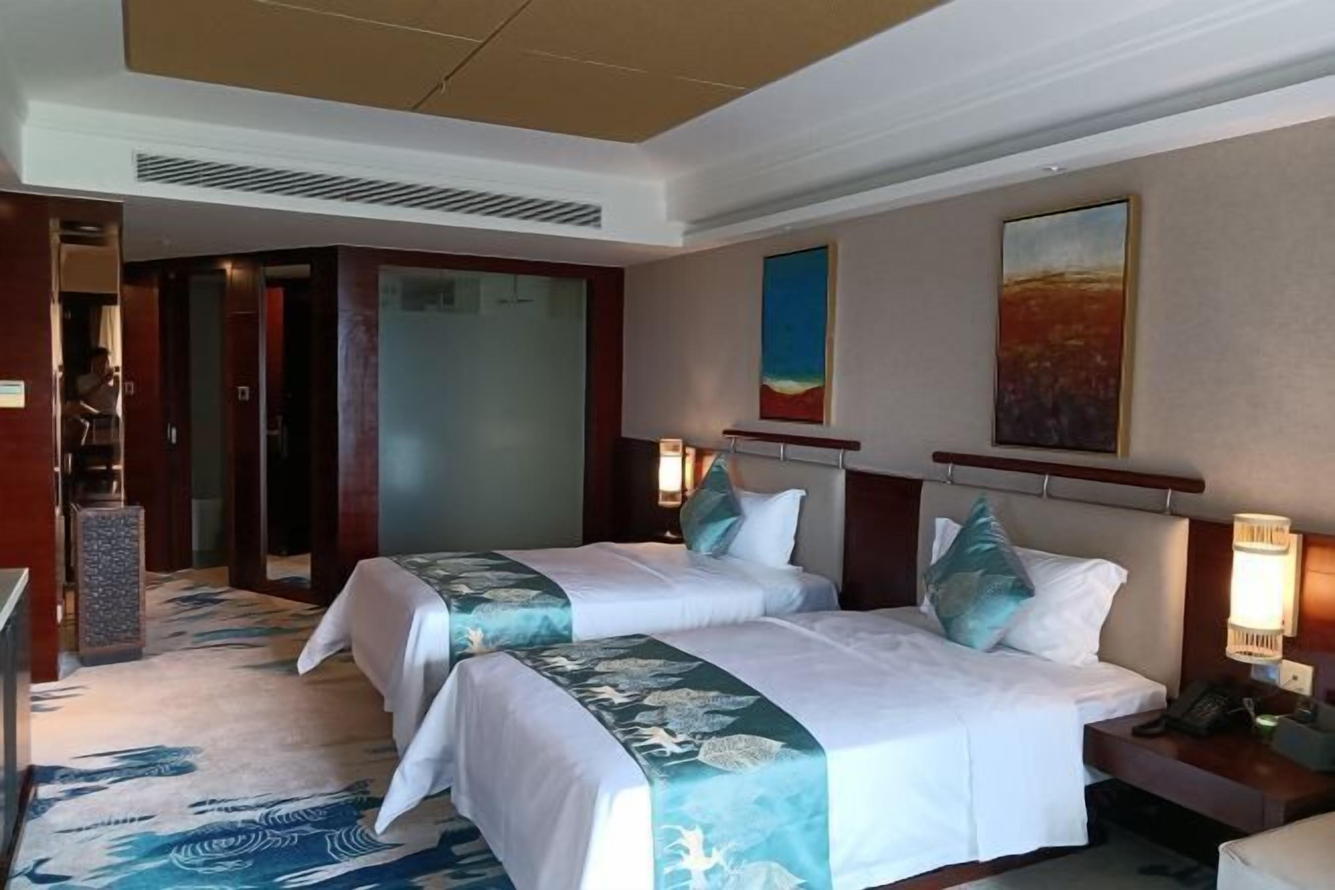 A hotel room with two beds, a desk, a chair, a nightstand, and a painting on the wall.