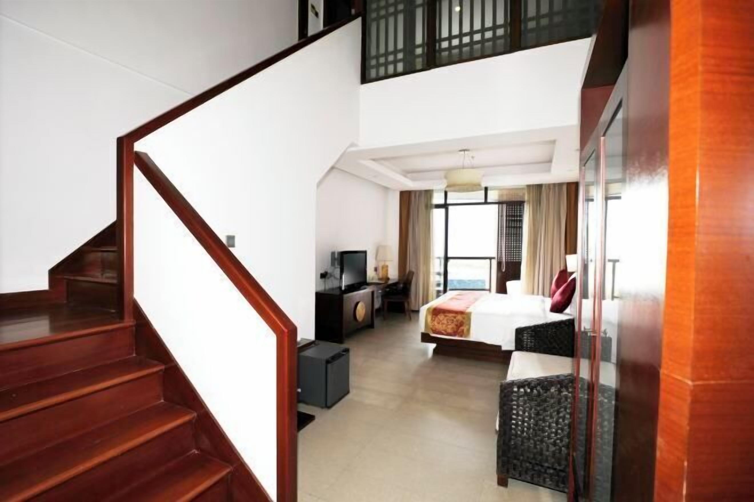 A hotel room with a wooden staircase, a bed with a red blanket, a desk with a chair, and a television.