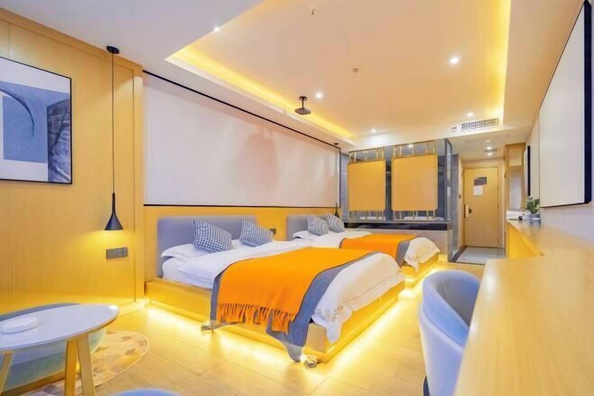 A modern hotel room with two beds, a small table, and a bathroom visible in the background.