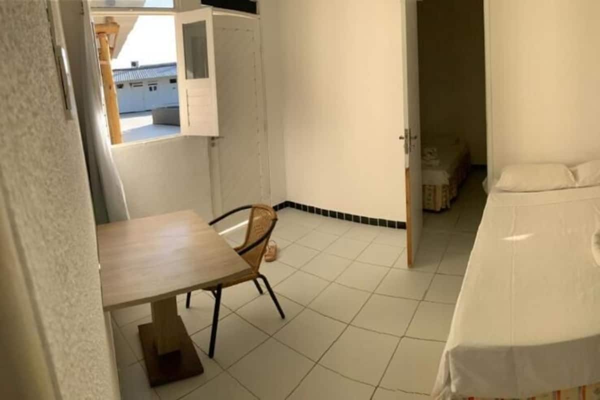 A small, clean room with a bed, a table, and a chair. There is a window providing natural light and a view of the outside.