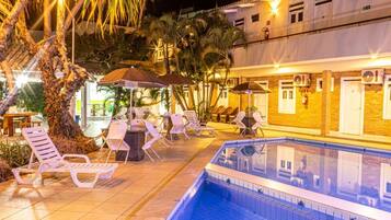 Zona de piscina con sillas de descanso y sombrillas, rodeada de palmeras y un edificio con unidades de aire acondicionado.