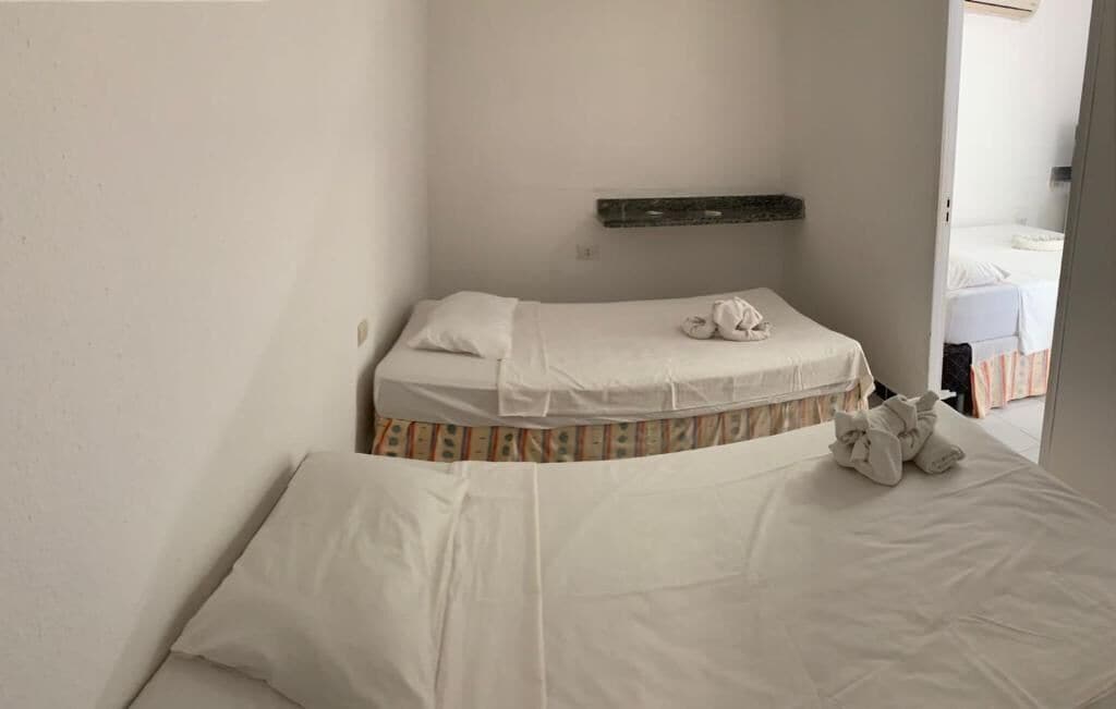 Quarto pequeno e simples com uma cama de solteiro, mesa de cabeceira e prateleira sobre a cama.