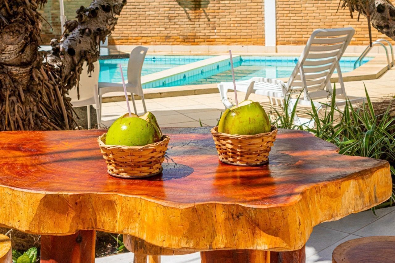 Une table en bois sur laquelle reposent deux paniers tressés remplis de jeunes noix de coco vertes, une piscine et, au fond, des chaises blanches.