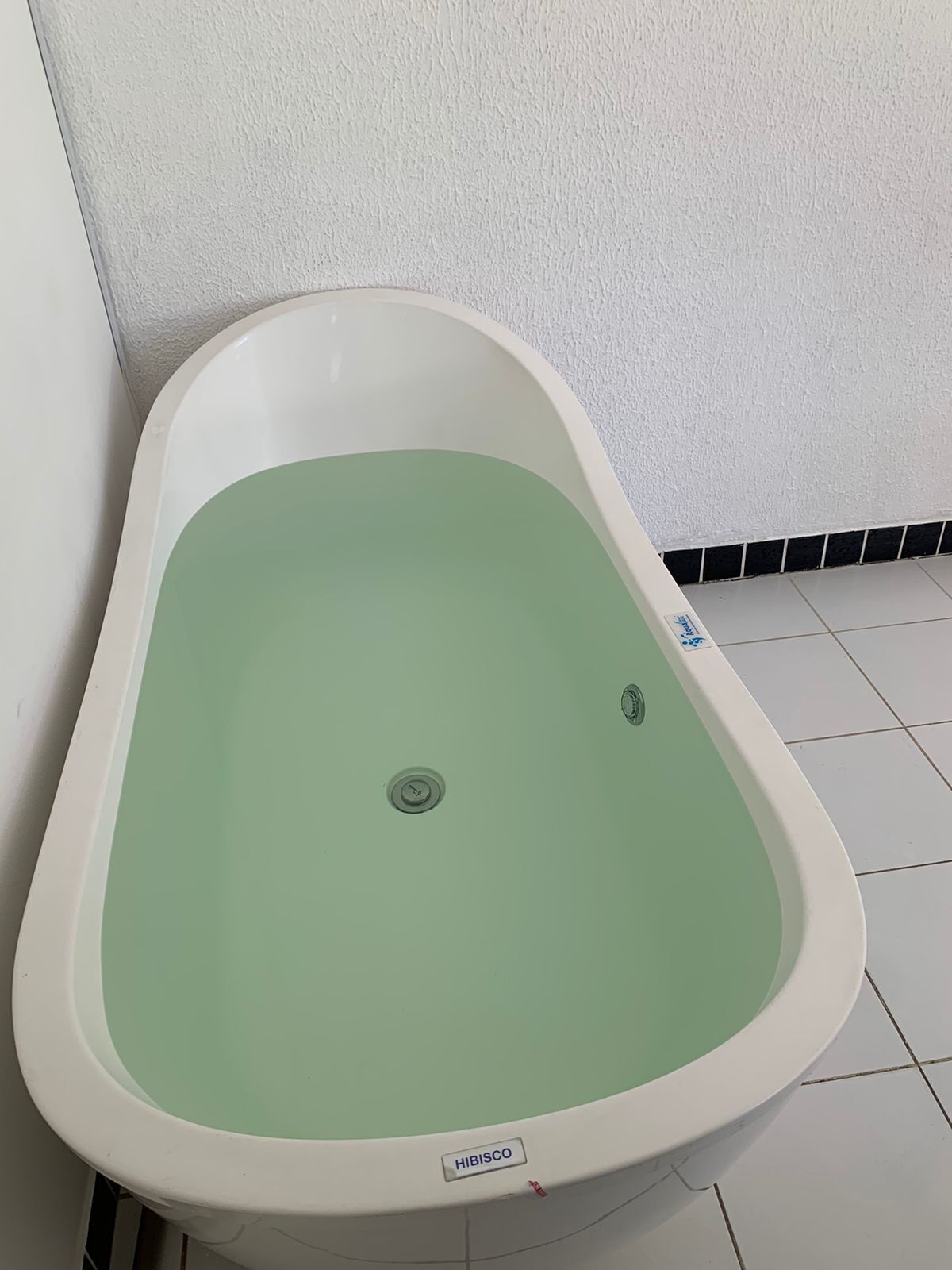 A white bathtub with a green interior, labeled "HIBISCO," in a corner of a room with white walls and tiled flooring.