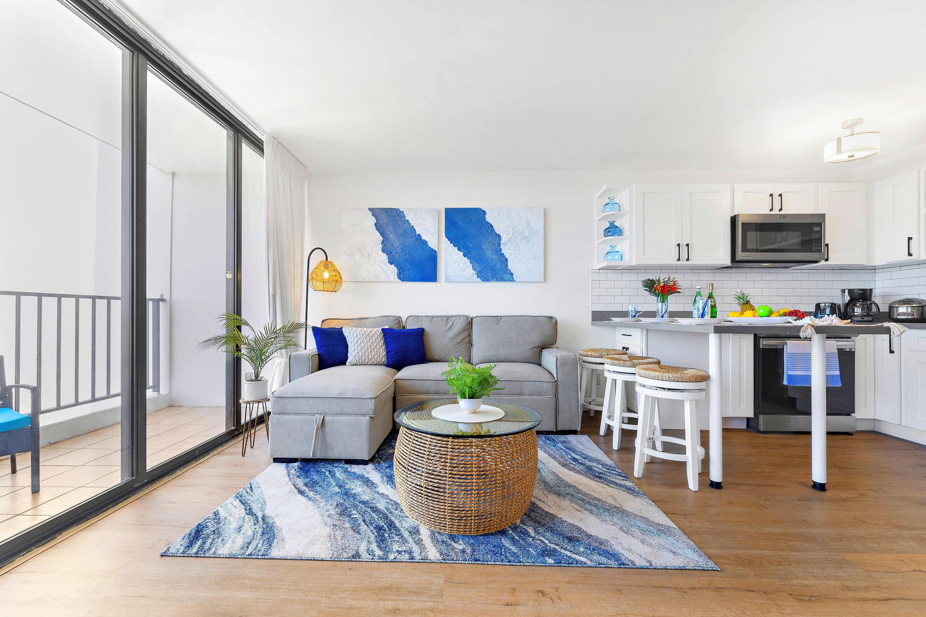 A modern apartment with a living area featuring a grey sofa, a round wicker coffee table, and a blue and white rug. The kitchen has white cabinets, a microwave, and a small island with stools.