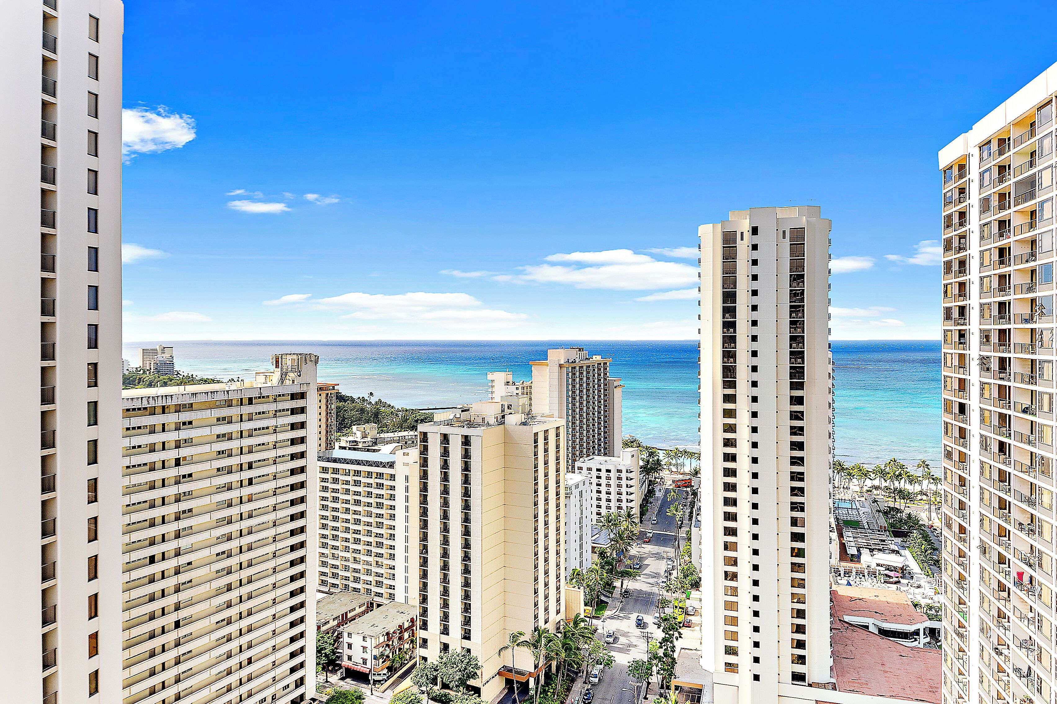 A high-rise hotel with multiple floors and balconies, overlooking a beach with clear blue water and palm trees.