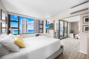 Un dormitorio con un ventanal grande, una cama, un escritorio y vistas de edificios altos y el océano.