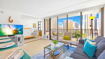 A modern living room with a sofa, a coffee table, and a TV. There is a balcony with a view of tall buildings.