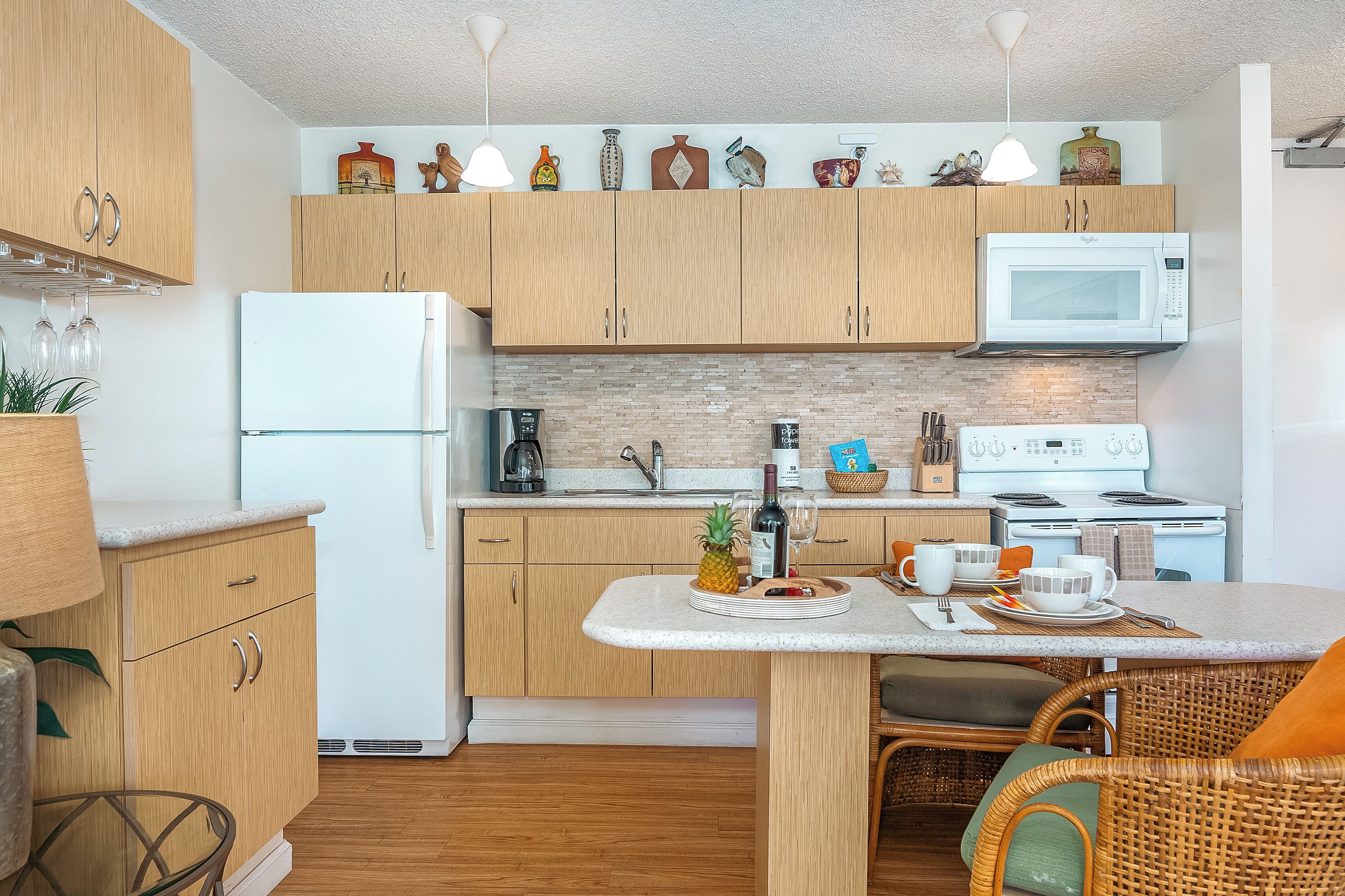 A modern kitchen with wooden cabinets, a white refrigerator, a microwave, and a stove. There is a dining table with chairs and a small island with a pineapple and wine glasses.