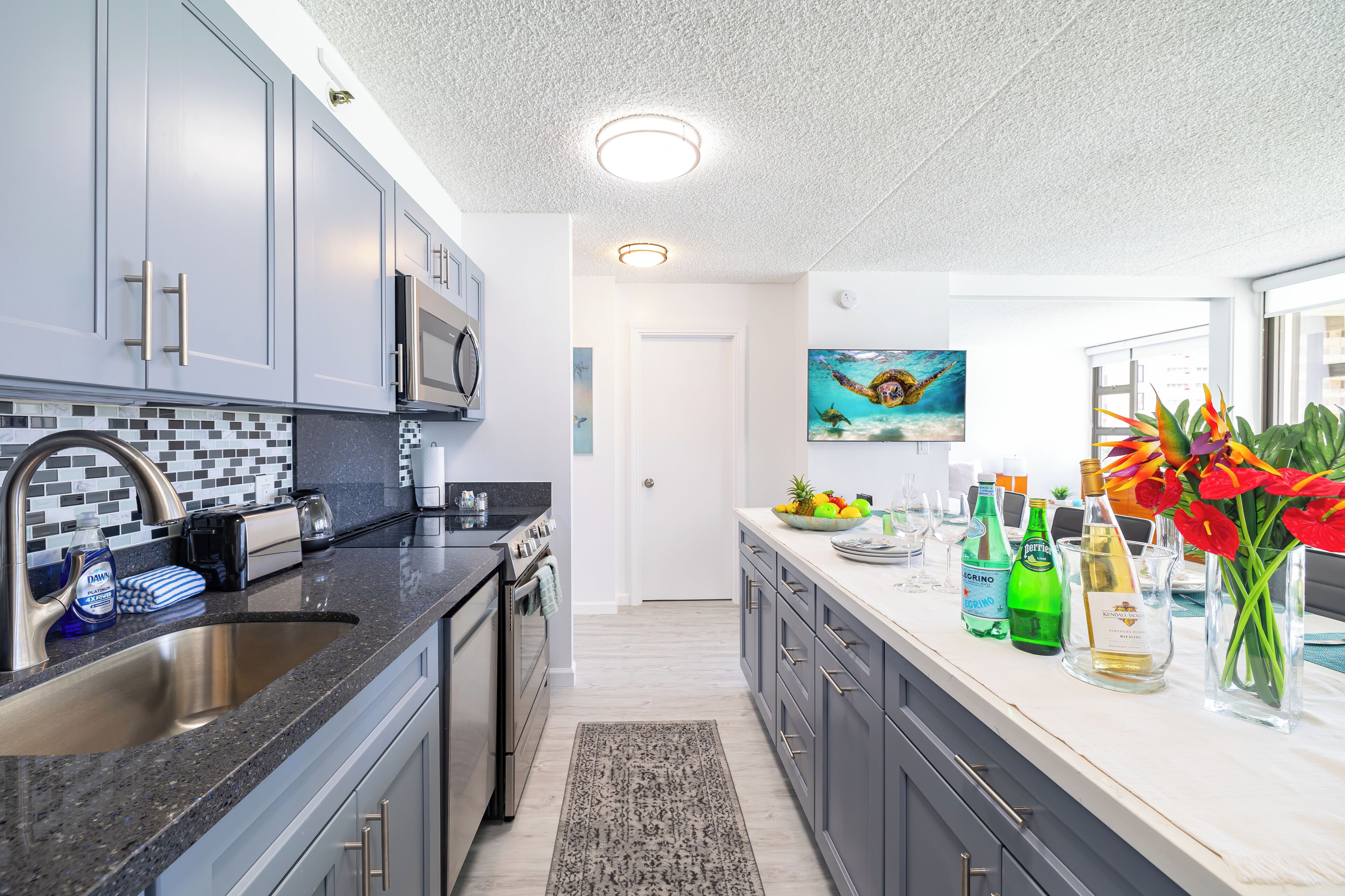 A modern kitchen with grey cabinets, a black countertop, and a tiled backsplash. There is a microwave above the stove, a sink with a faucet, and a rug on the floor.