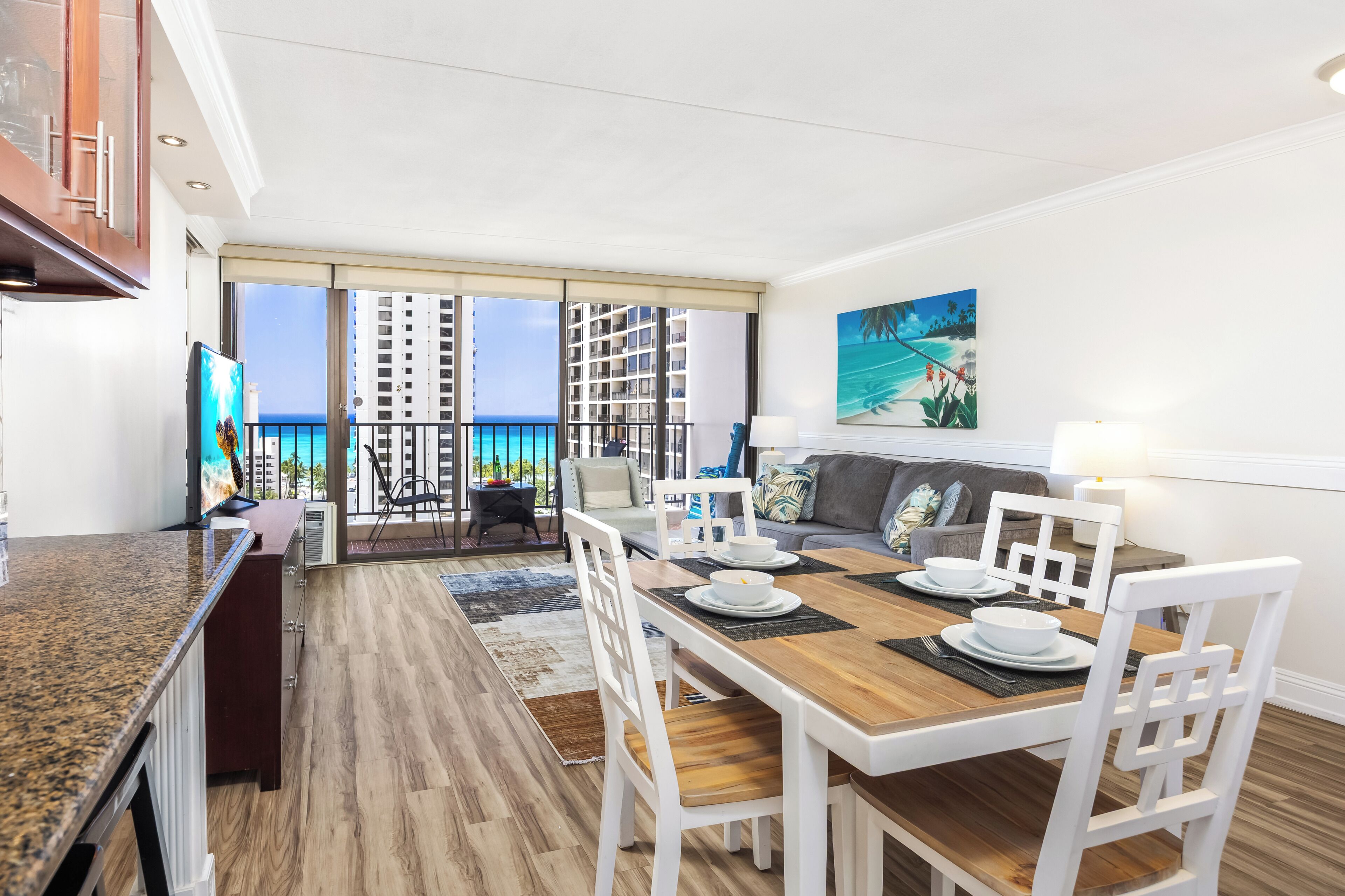 A modern dining area with a wooden table and white chairs, a kitchen with granite countertops, and a balcony with a view of the ocean and high-rise buildings.